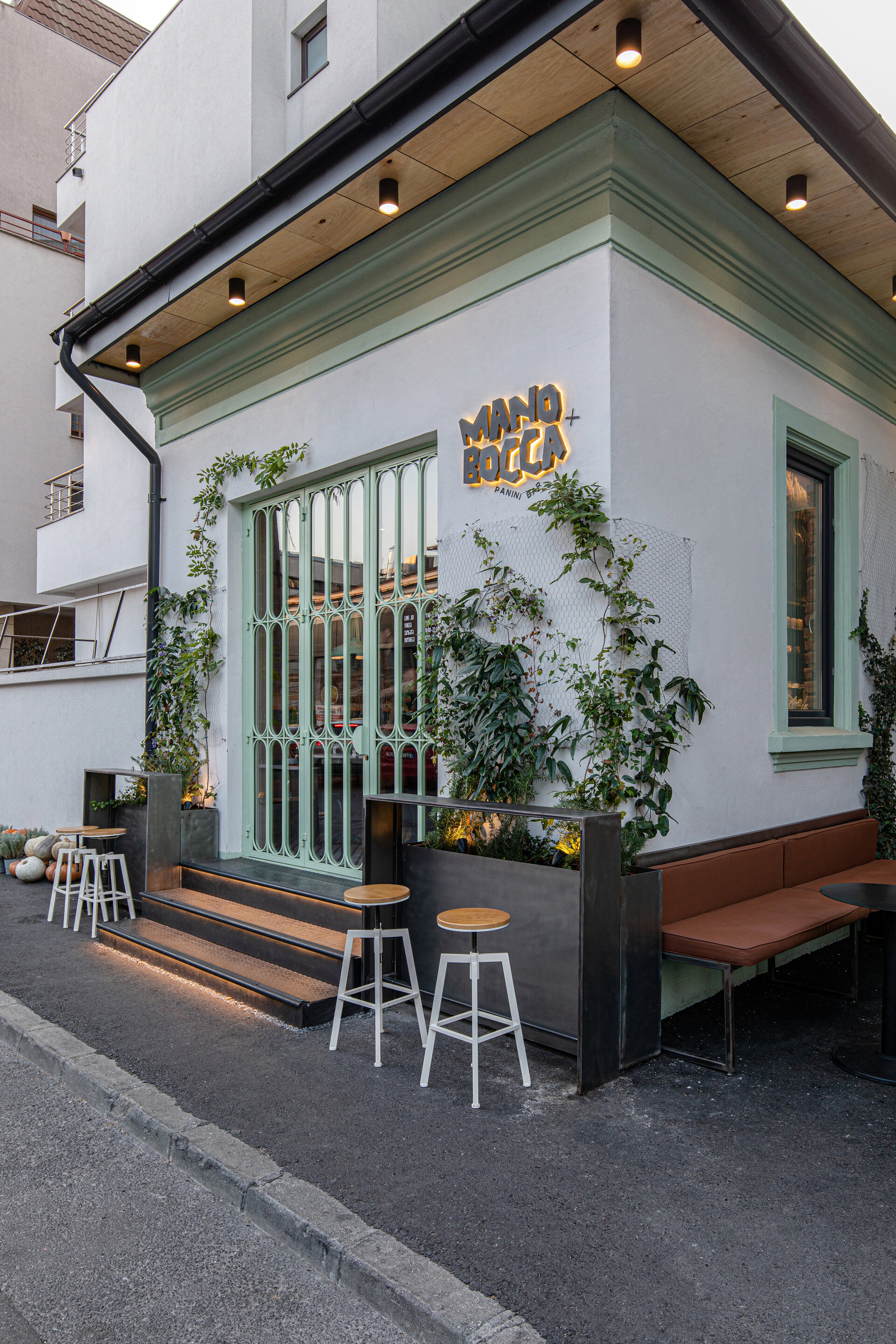Storefront Mano+Bocca Panini Bar — fațadă cu logo brushed, geam de fier verde-mentă, planters cu verdeață
